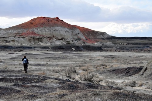 A lone hiker walks through a barren landscape with colorful hills under a cloudy sky.