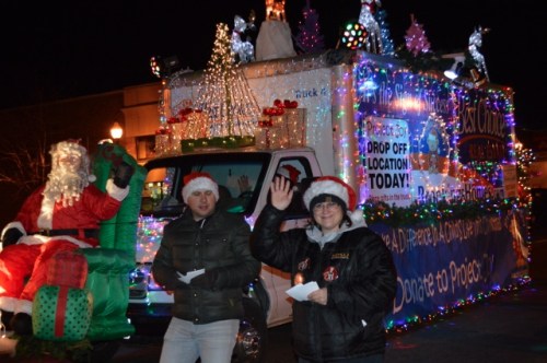 A festive parade float decorated with lights, featuring Santa and two people in holiday attire waving to the crowd.