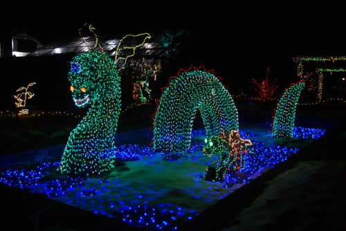 A colorful light display featuring a dragon-like creature emerging from a blue-lit pond, surrounded by festive decorations.