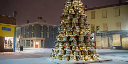 A festive Christmas tree made of golden bells, adorned with red ribbons, in a snowy urban setting at night.