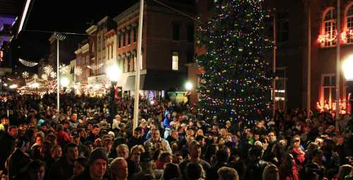 A large crowd gathers around a brightly lit Christmas tree in a festive downtown area at night.