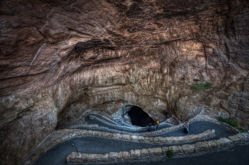 A winding stone path leads into a dark cave, surrounded by textured rock formations and sparse vegetation.