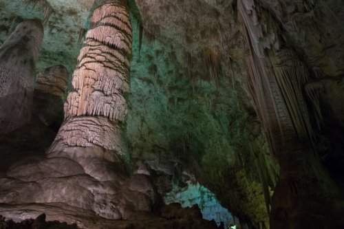A stunning cave interior featuring tall, intricately formed stalagmites and colorful lighting illuminating the rock formations.