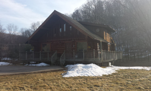 A large log cabin with a wraparound porch, surrounded by snow and grass, set against a wooded hillside.