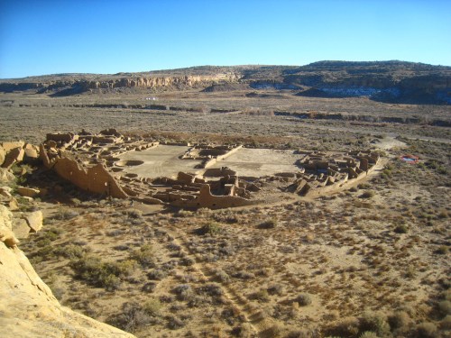 Ancient ruins of a circular structure in a desert landscape, surrounded by rocky hills and sparse vegetation.