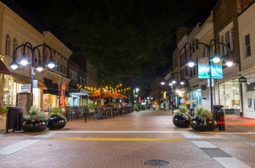 A quiet, illuminated street at night with outdoor seating, planters, and shops lining the walkway.