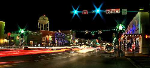 A festive street scene at night, adorned with colorful holiday lights and traffic moving through the illuminated area.