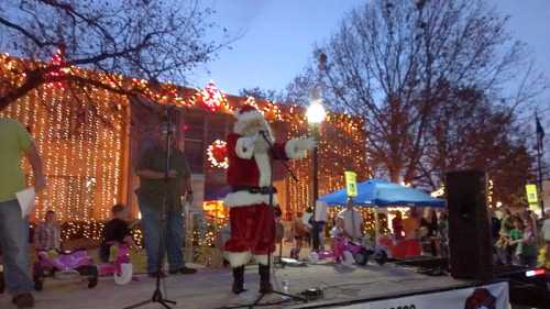 A man dressed as Santa Claus stands on stage, holding a lantern, with festive lights and decorations in the background.