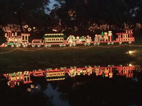 Colorful holiday lights depicting a train and festive scenes, reflected in a calm body of water.