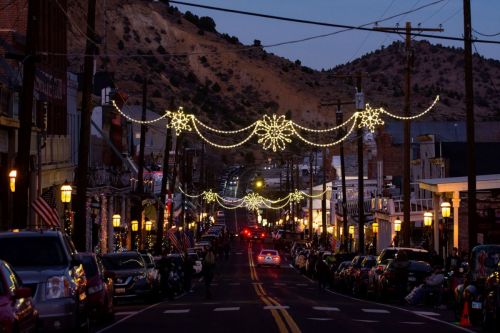 A festive street scene at dusk, adorned with holiday lights and decorations, lined with parked cars and mountains in the background.