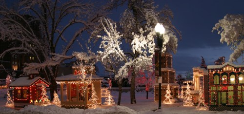 A snowy scene featuring colorful holiday lights on small buildings and trees, creating a festive winter atmosphere.