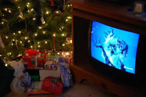 A vintage TV shows a scene from a holiday movie, surrounded by wrapped gifts and a decorated Christmas tree.