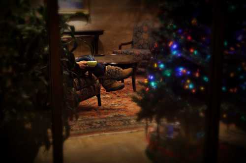 A cozy scene with a person sitting in a chair, holding a cup, next to a decorated Christmas tree.