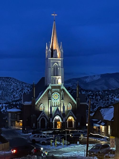 A lit church with a tall steeple and stained glass windows, surrounded by snow-covered mountains at dusk.