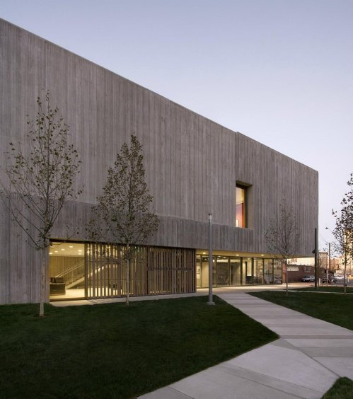 Modern concrete building with large windows, surrounded by grass and small trees, under a twilight sky.