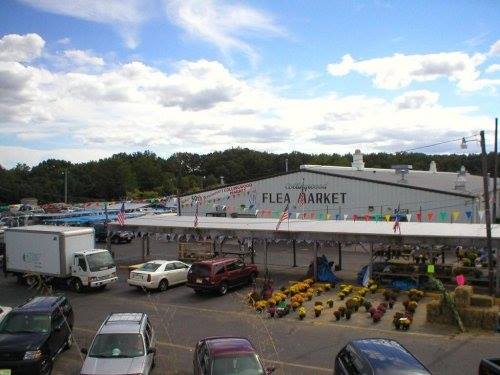 A bustling flea market with colorful banners, parked cars, and vendors under a partly cloudy sky.