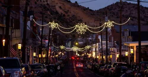 A festive street scene at night, adorned with snowflake lights and lined with parked cars, set against a mountainous backdrop.