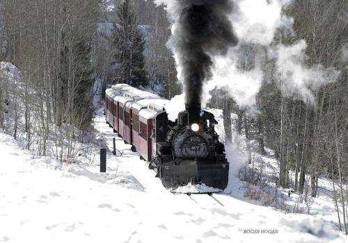 A vintage steam train chugs through a snowy landscape, billowing smoke against a backdrop of trees.