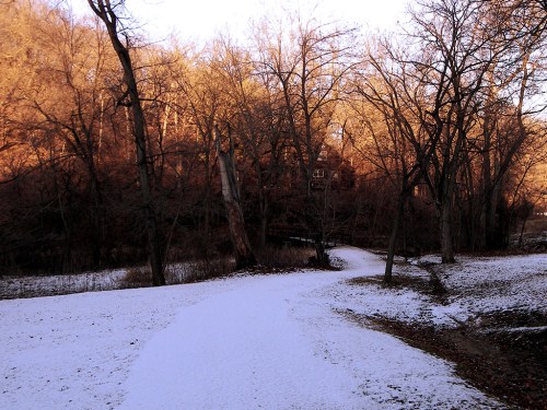 A snowy path winds through a wooded area with bare trees and a warm glow from the setting sun in the background.