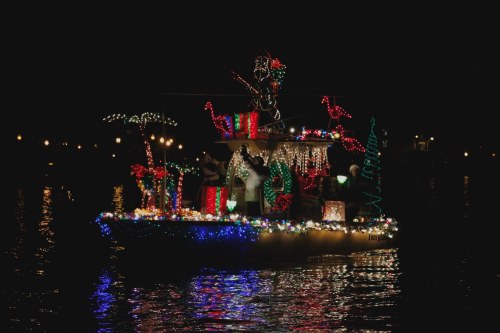 A brightly lit boat decorated with festive lights and holiday decorations glides across a dark, reflective water surface.