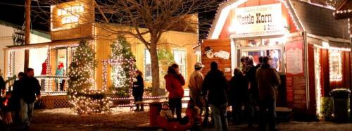 A festive scene with people gathered outside a brightly lit building, decorated for the holidays with lights and trees.