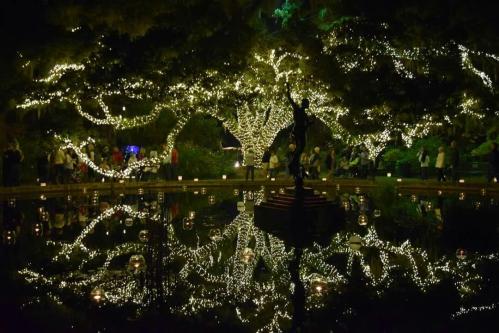 A beautifully lit garden at night, featuring a statue and reflections in a calm pond surrounded by festive lights.