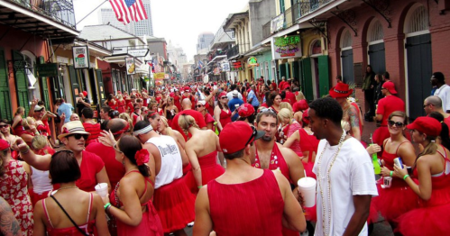 A lively crowd in red outfits celebrates in a festive street, with buildings and American flags in the background.