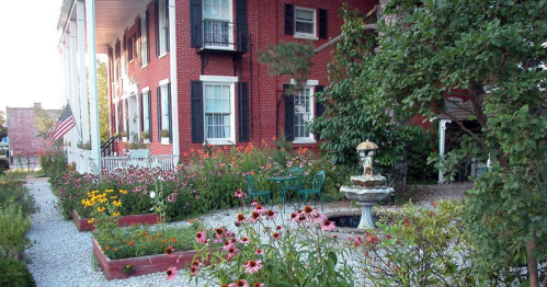 A charming brick house with a garden featuring colorful flowers and a fountain, surrounded by gravel pathways.