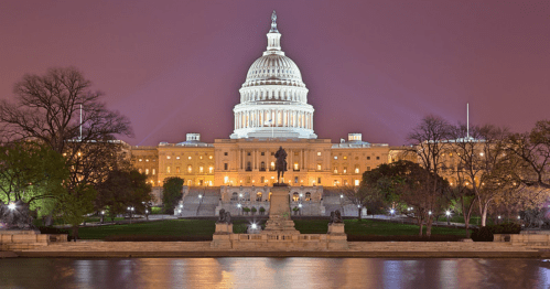 The U.S. Capitol building illuminated at night, reflecting in the water with trees in the foreground.