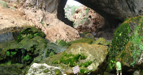 A child stands on rocky terrain beneath a large natural arch, surrounded by moss-covered boulders and rugged cliffs.