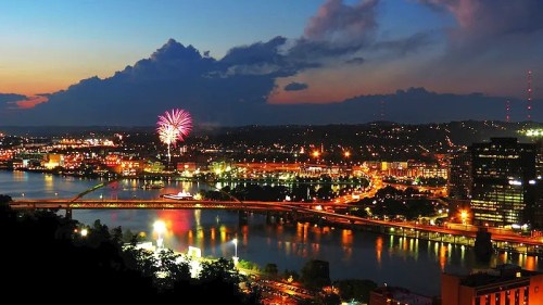 A vibrant city skyline at dusk, with fireworks illuminating the sky over a river and bridges.