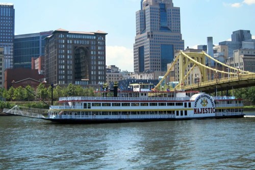 A large riverboat named "Majestic" sails on a river with city buildings and a yellow bridge in the background.