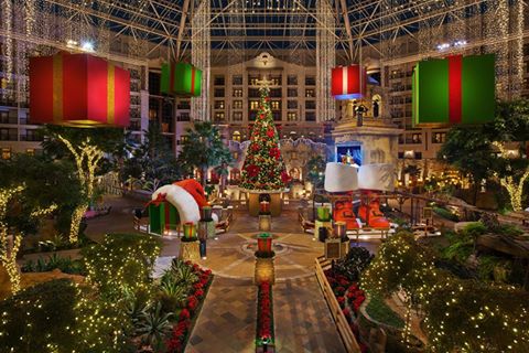 A festive indoor scene featuring a large Christmas tree, colorful gift boxes, and holiday decorations in a beautifully lit atrium.
