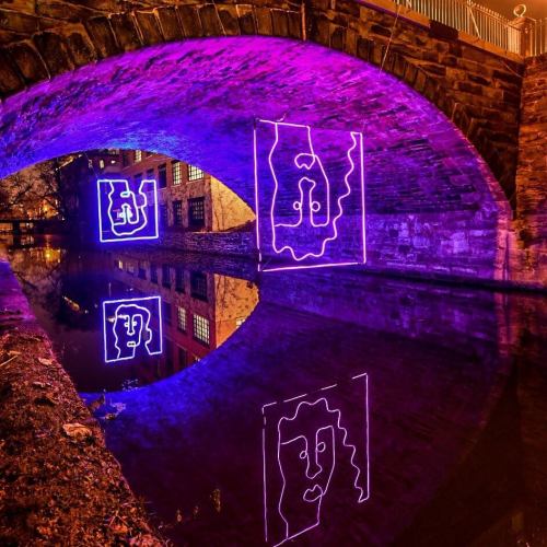 A stone bridge arching over water, illuminated by purple light reflections and geometric shapes on the surface.