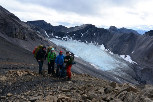 A group of four hikers stands on a rocky mountain trail with a glacier and rugged peaks in the background.