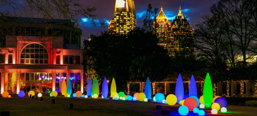 Colorful illuminated spheres and cones in a park at night, with city skyscrapers glowing in the background.