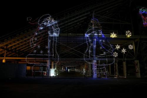 Colorful holiday lights in the shape of bells and figures decorate a dark, open space, creating a festive atmosphere.