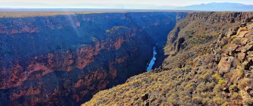 A panoramic view of a deep canyon with a winding river at the bottom, surrounded by rugged cliffs and sparse vegetation.