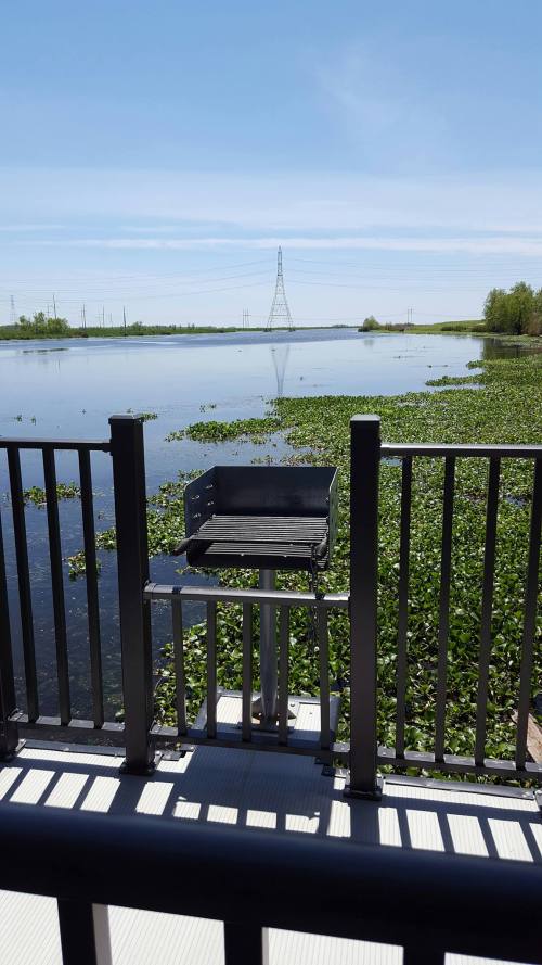 A grill on a deck overlooking a calm lake with greenery and power lines in the background under a clear blue sky.