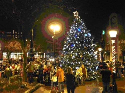 A festive crowd gathers around a brightly lit Christmas tree at night, with streetlights illuminating the scene.