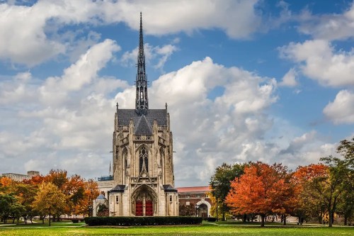 Gothic-style church with a tall spire, surrounded by colorful autumn trees under a partly cloudy sky.