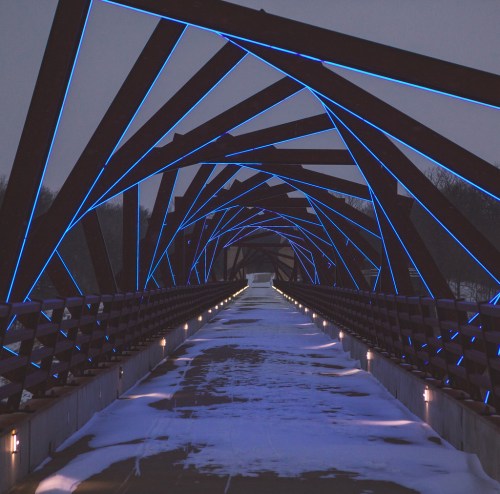 A snow-covered bridge with blue LED lights illuminating its geometric structure at dusk.