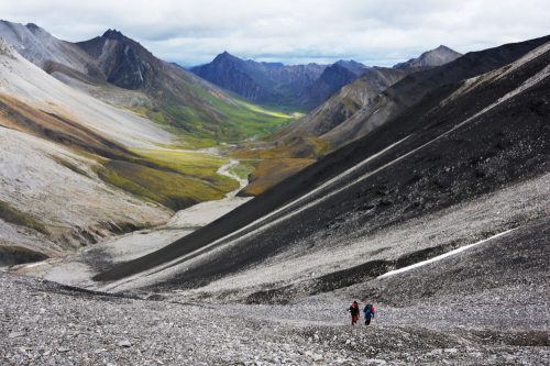 Two hikers trek down a rocky slope in a vast mountain valley, surrounded by rugged peaks and lush green fields.