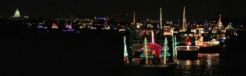 Colorful boats adorned with festive lights on a dark river, with a city skyline in the background.