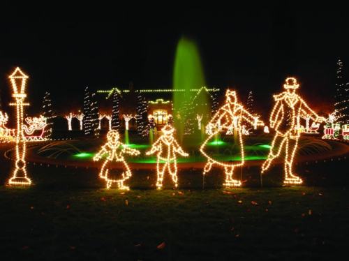 Illuminated figures in festive attire stand by a fountain, surrounded by twinkling holiday lights at night.