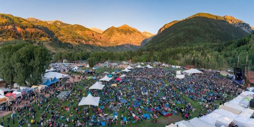 Aerial view of a large outdoor festival with crowds, tents, and mountains in the background during sunset.