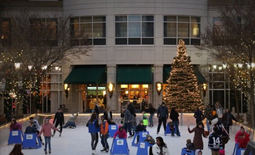 People ice skating in a festive outdoor rink with a Christmas tree and holiday lights in the background.