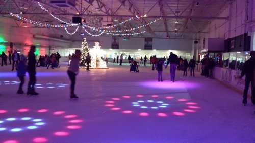 A lively ice skating rink decorated with colorful lights and a Christmas tree, filled with skaters enjoying the scene.
