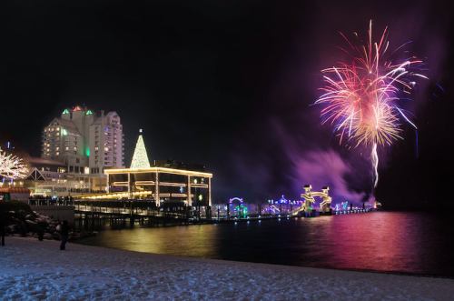Colorful fireworks illuminate the night sky over a festive waterfront with holiday lights and a snowy foreground.