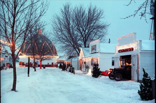 A snowy street scene with decorated buildings and trees, illuminated by festive lights during winter.